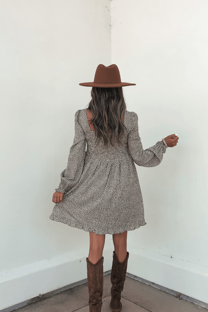 A woman in a brown hat and boots stands facing a white wall, holding her Olive Floral Print Ruched Mini Dress - FINAL SALE.