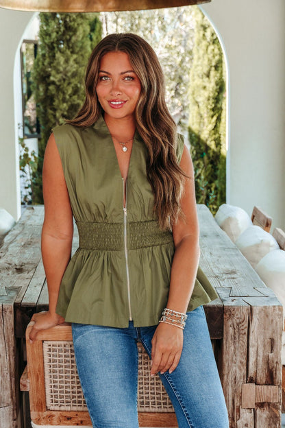 Woman with long brown hair in the Olive Grove Smocked Peplum Top and jeans by a rustic table—perfect for your fall wardrobe.