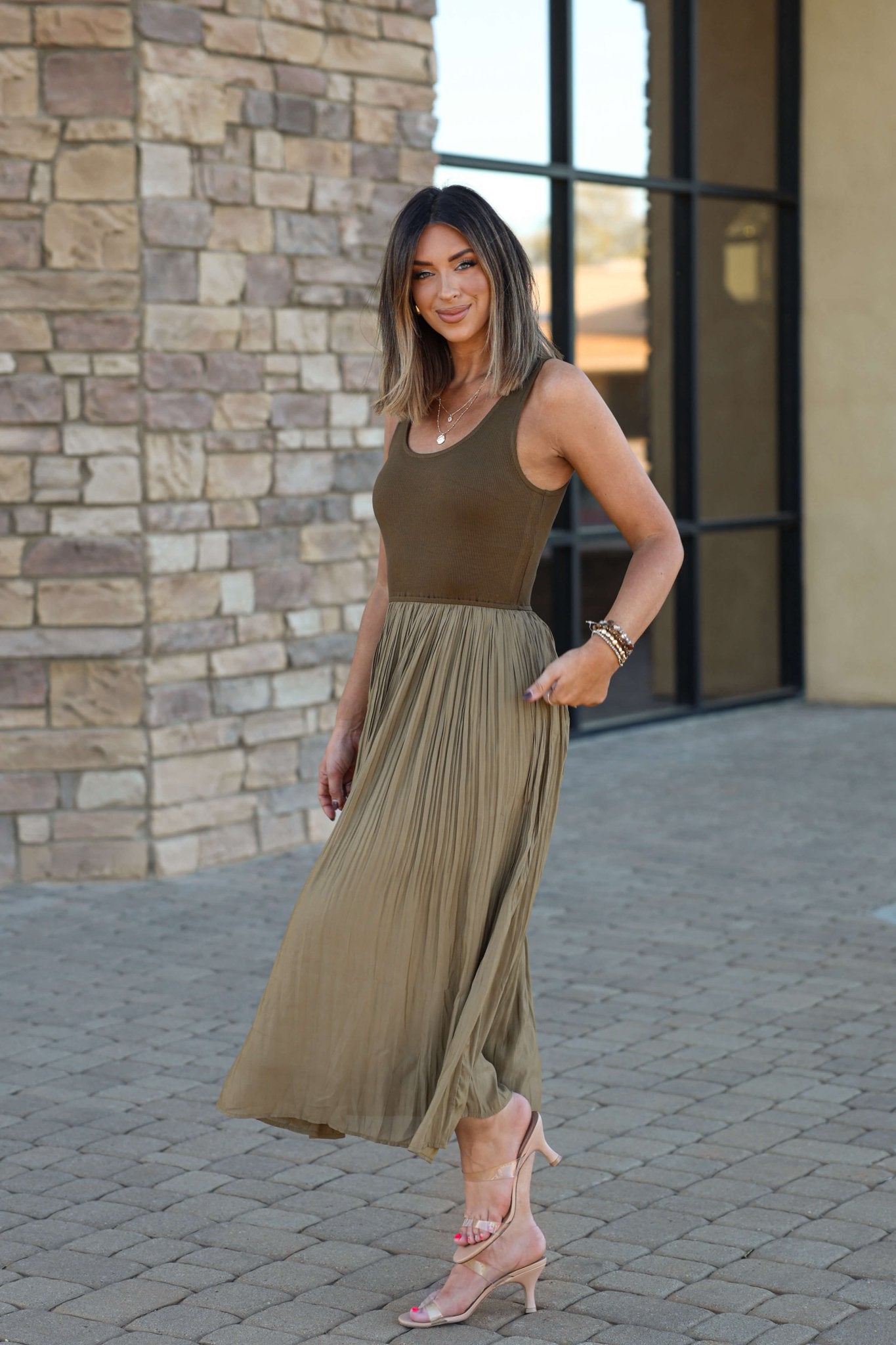 Woman smiles near a stone building in the Olive Scoop Neck Pleated Tank Midi Dress - DOORBUSTER and pink heels.