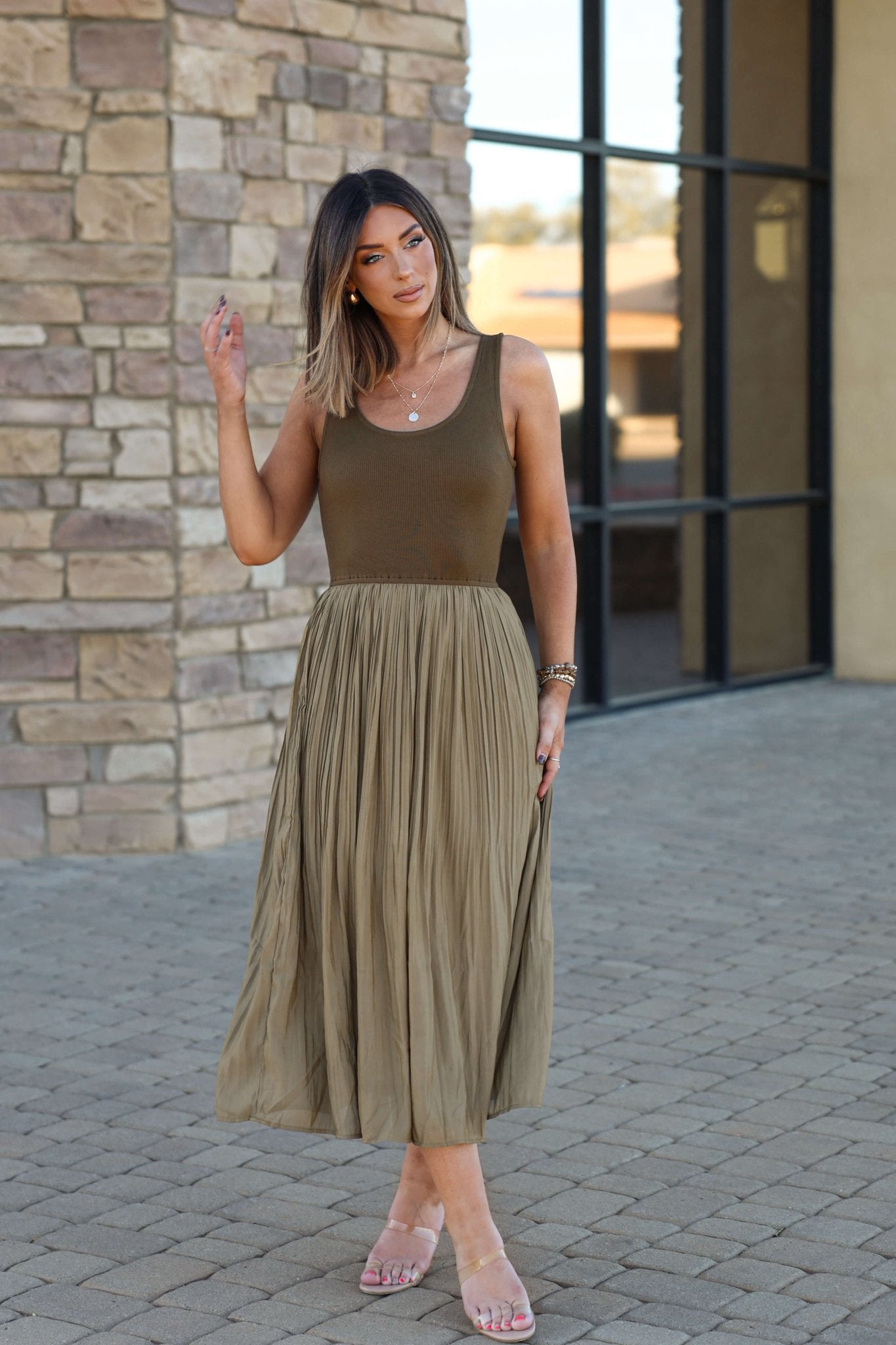 Woman wearing the Olive Scoop Neck Pleated Tank Midi Dress, standing on a paved path by a modern stone and glass building.
