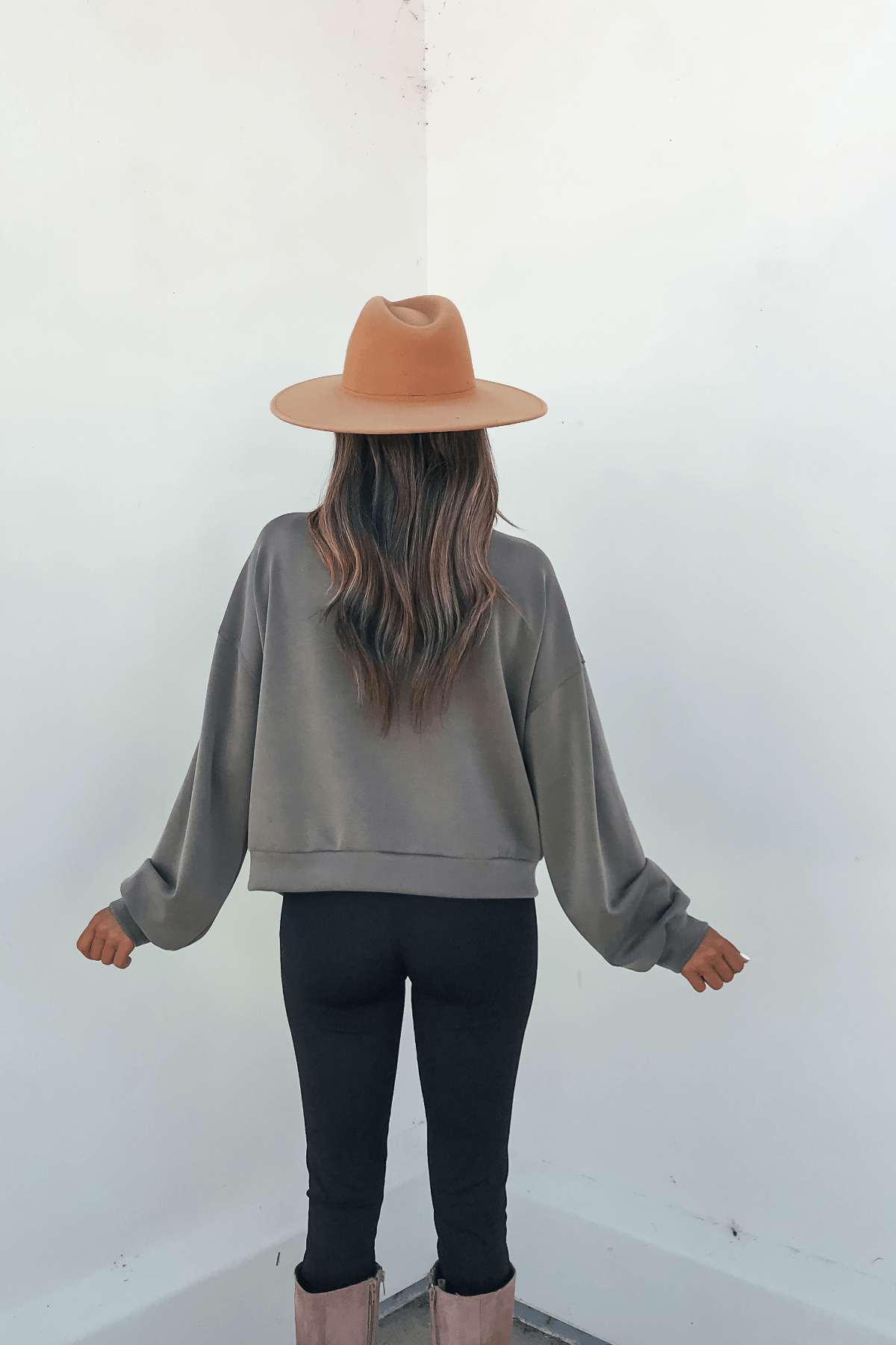 Wearing the Olive Soft Cloud Scuba Top, tan hat, gray sweater, and boots, someone stands facing a white wall—everyday essentials.