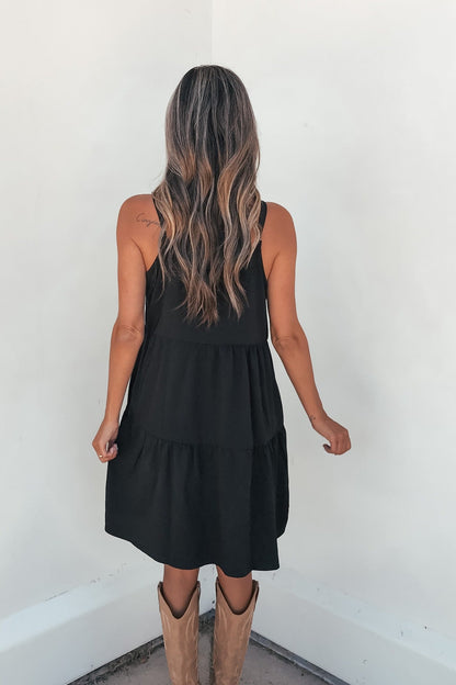 Woman with long wavy hair wears the On Point Tiered Mini Dress - Black, facing a white wall, back to camera.