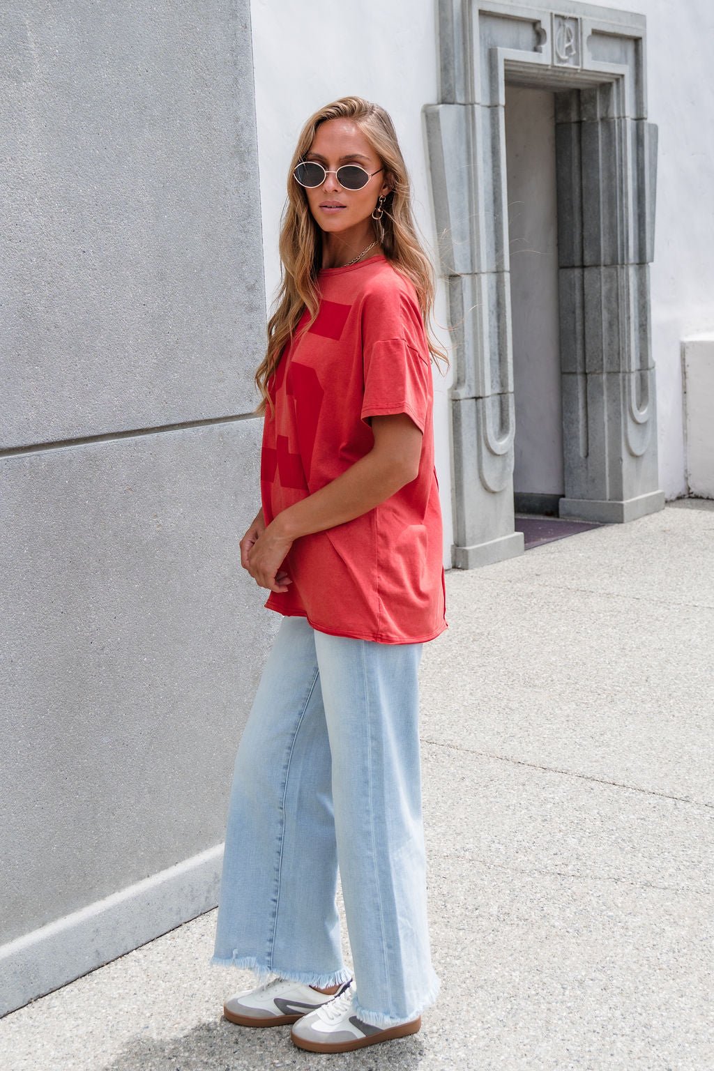 Woman in sunglasses wears an Oversized Graphic Tee - Red with light blue wide-leg jeans and white sneakers near a stone building.