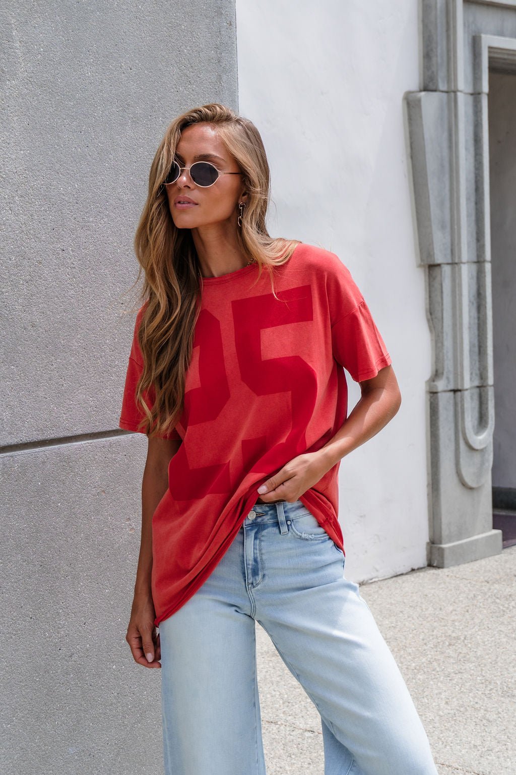 Woman in sunglasses wears the Oversized Graphic Tee - Red and light blue jeans, leaning against a wall outdoors.