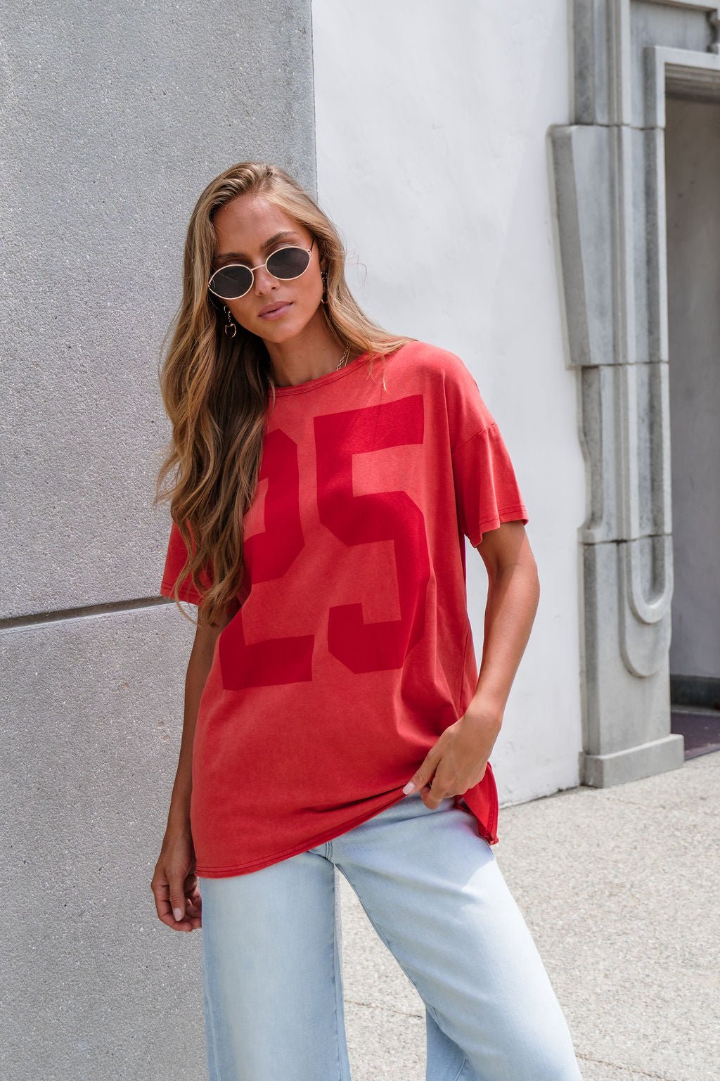 Woman in sunglasses wearing an Oversized Graphic Tee - Red and light blue jeans by a light-colored building.