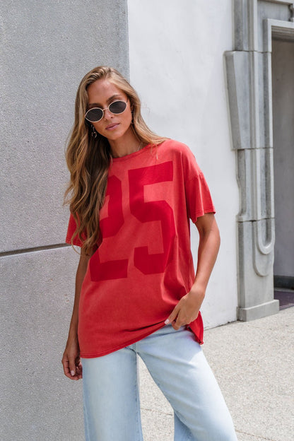 Woman in sunglasses wearing an Oversized Graphic Tee - Red and light blue jeans by a light-colored building.