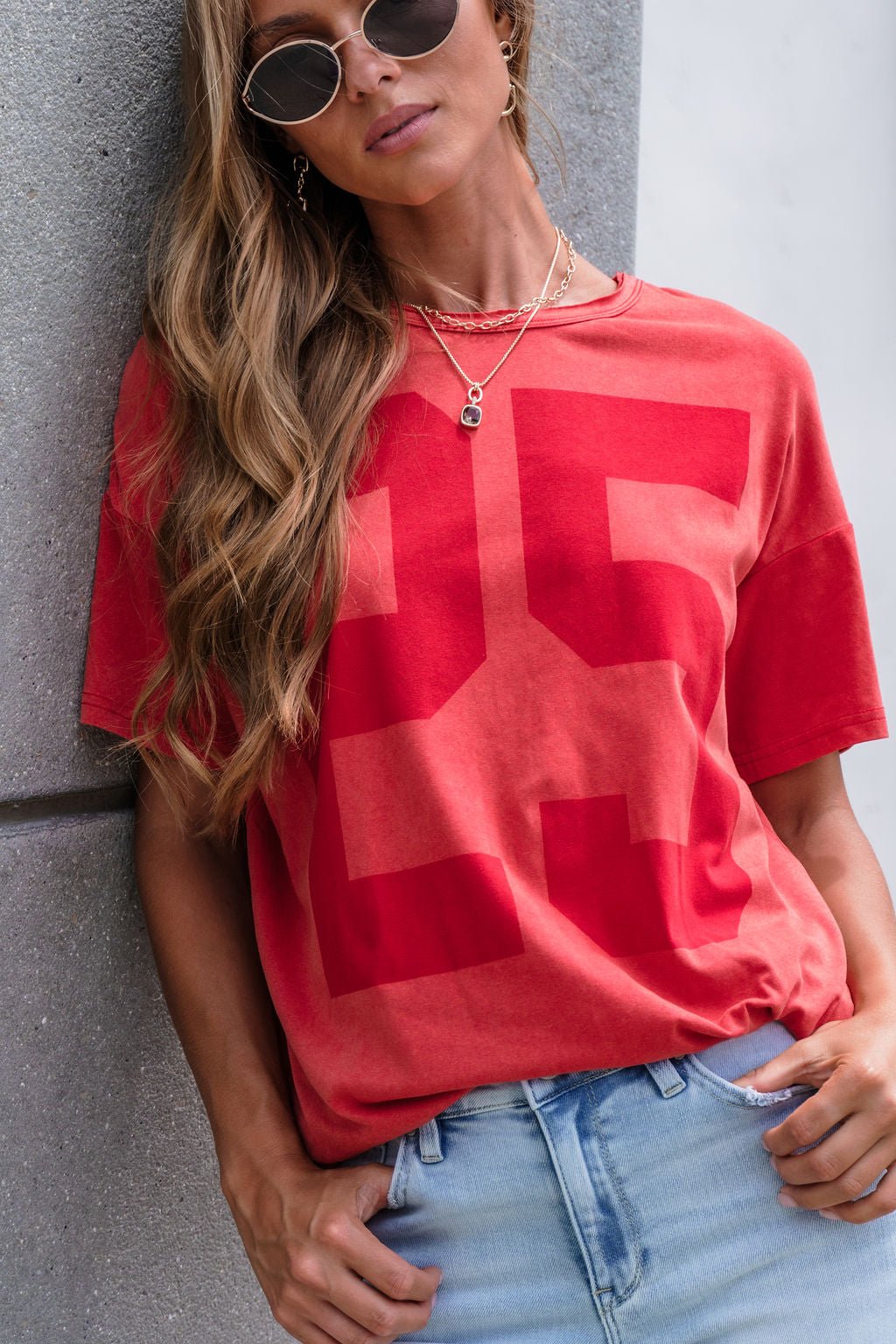 Woman in an Oversized Graphic Tee - Red, layered necklaces, and light blue jeans leans on a textured gray wall wearing cool shades.