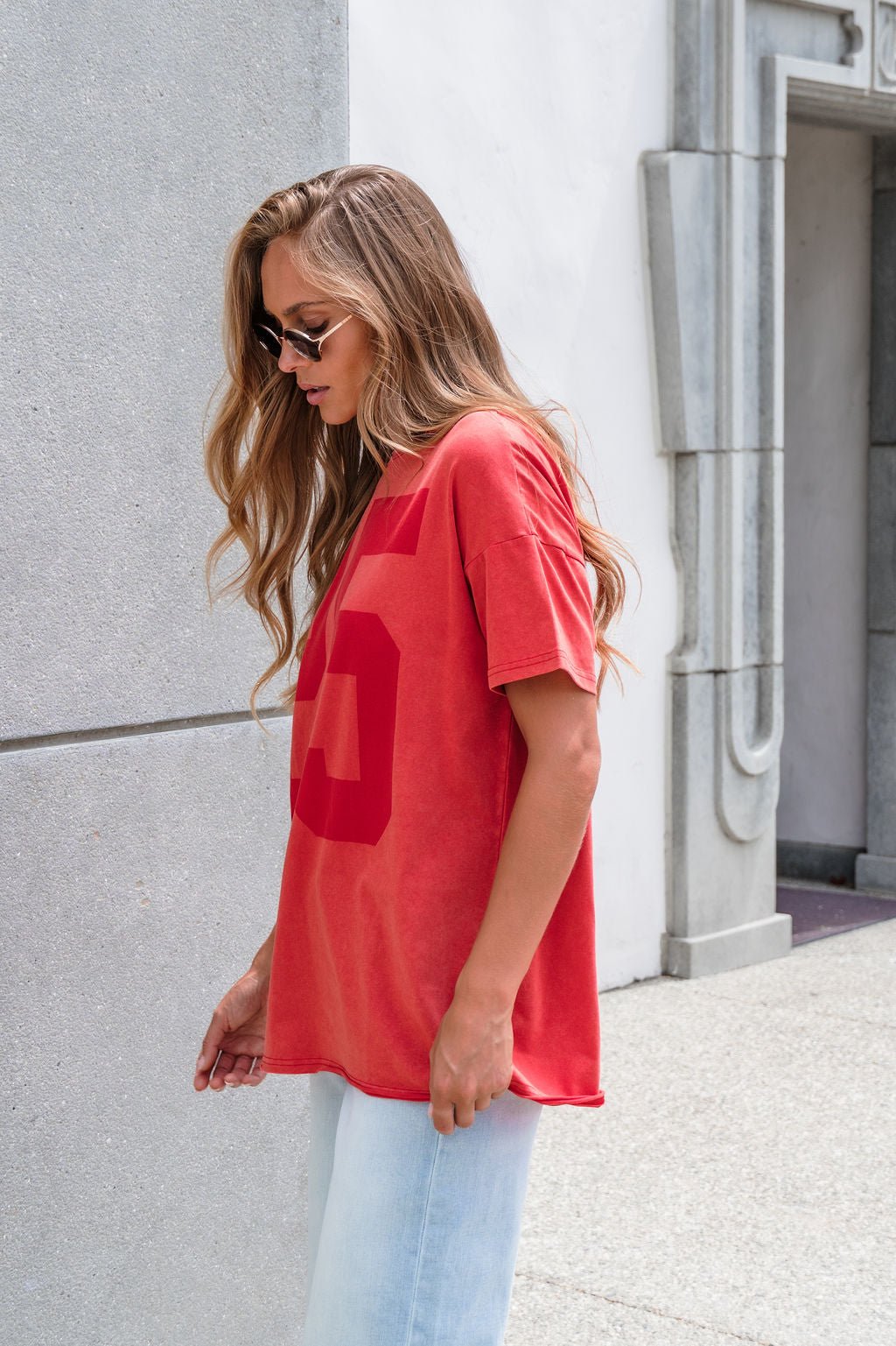 Woman with long hair wears sunglasses, an Oversized Graphic Tee - Red, and light jeans by a concrete wall outside a building.