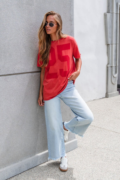 Woman in sunglasses leans against a wall, wearing an Oversized Graphic Tee - Red, wide-leg jeans, and white sneakers.