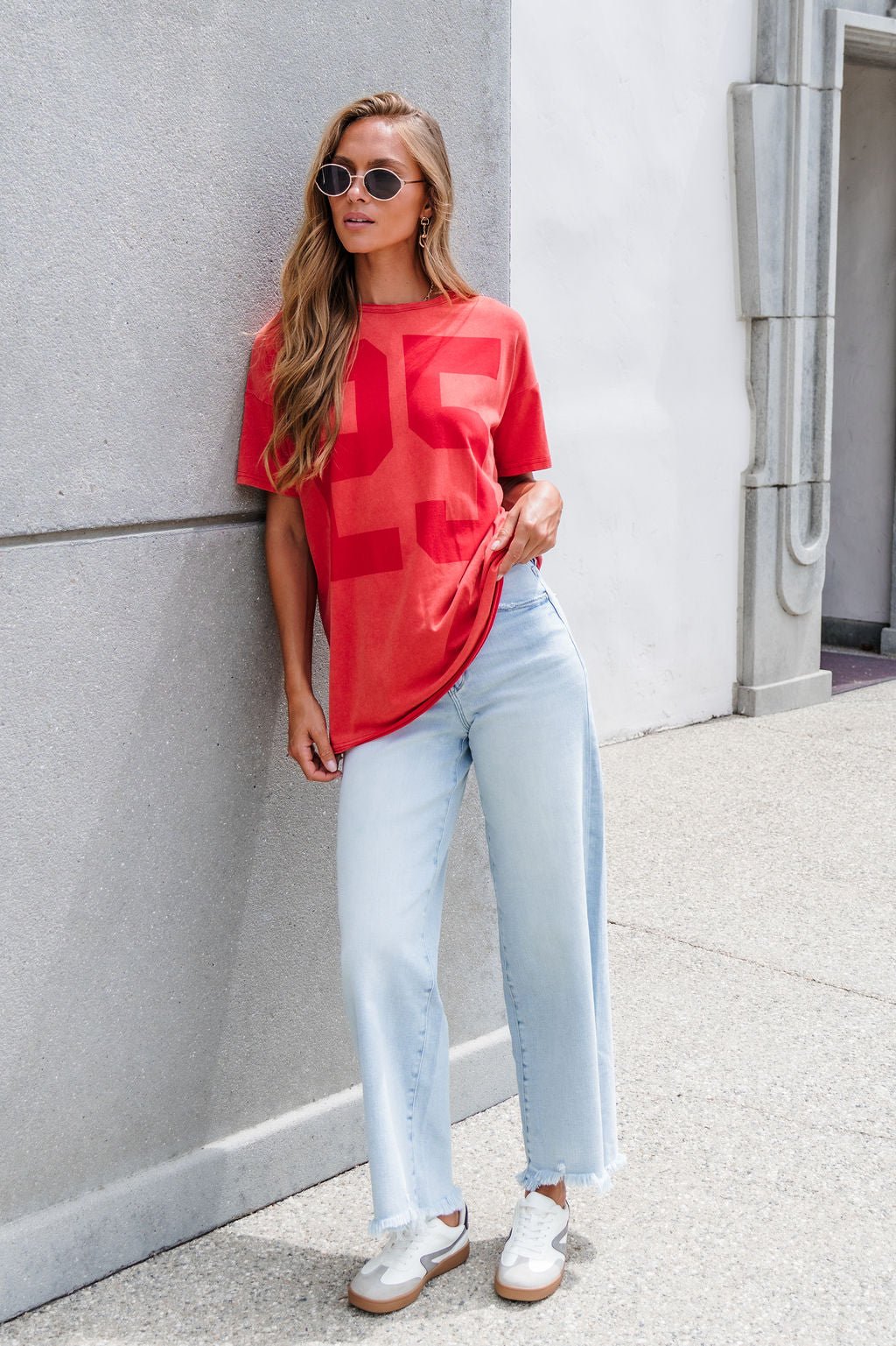 Woman in sunglasses leans on a wall, wearing an Oversized Graphic Tee - Red, light blue jeans, and white sneakers.