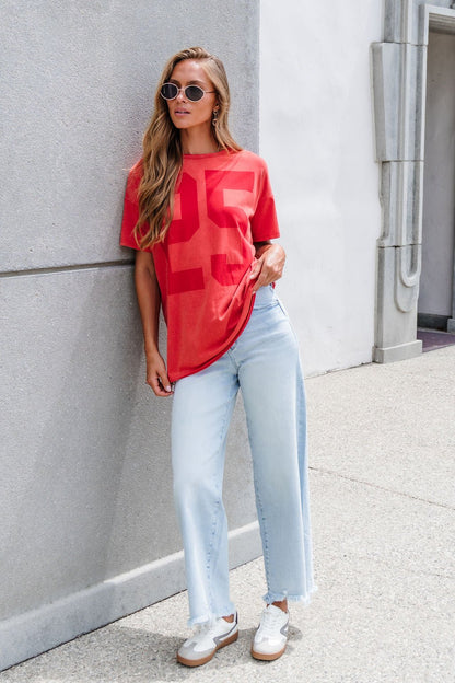 Woman in sunglasses leans on a wall, wearing an Oversized Graphic Tee - Red, light blue jeans, and white sneakers.