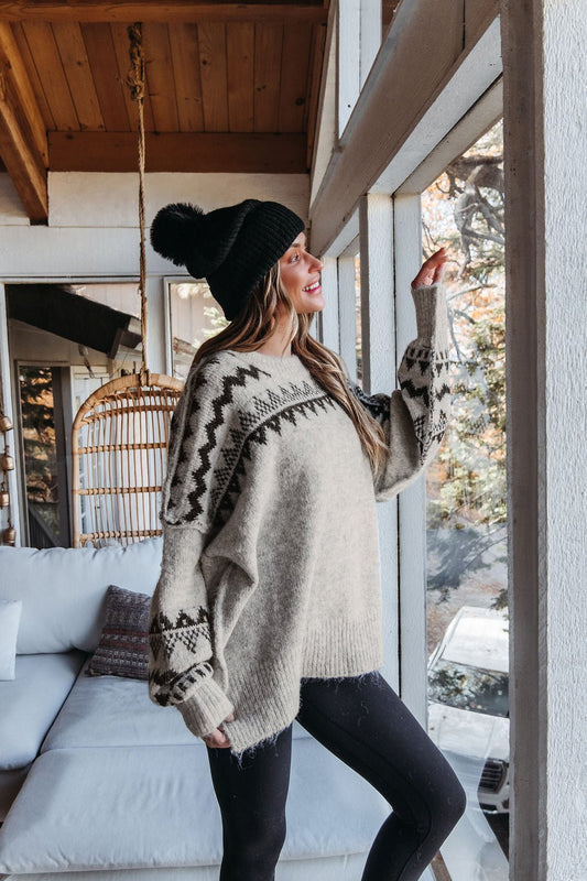 Woman in an Oversized Mocha Fair Isle Pullover Sweater stands by a window, smiling as she looks outside.