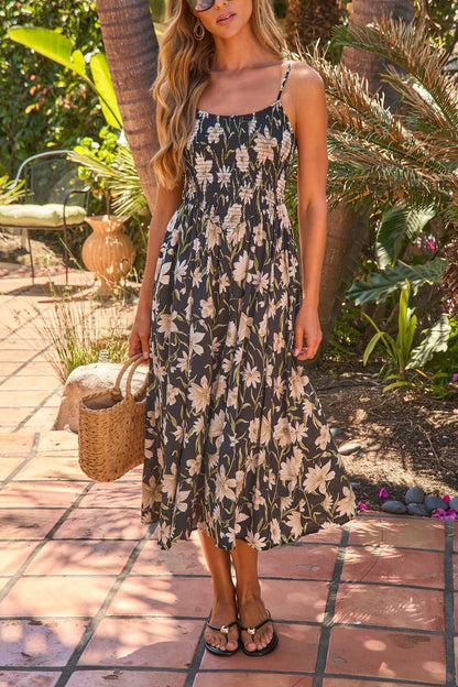 Woman in the Palm Shade Floral Midi Dress - Black, holding a straw bag on a sunlit patio with green plants in the background.