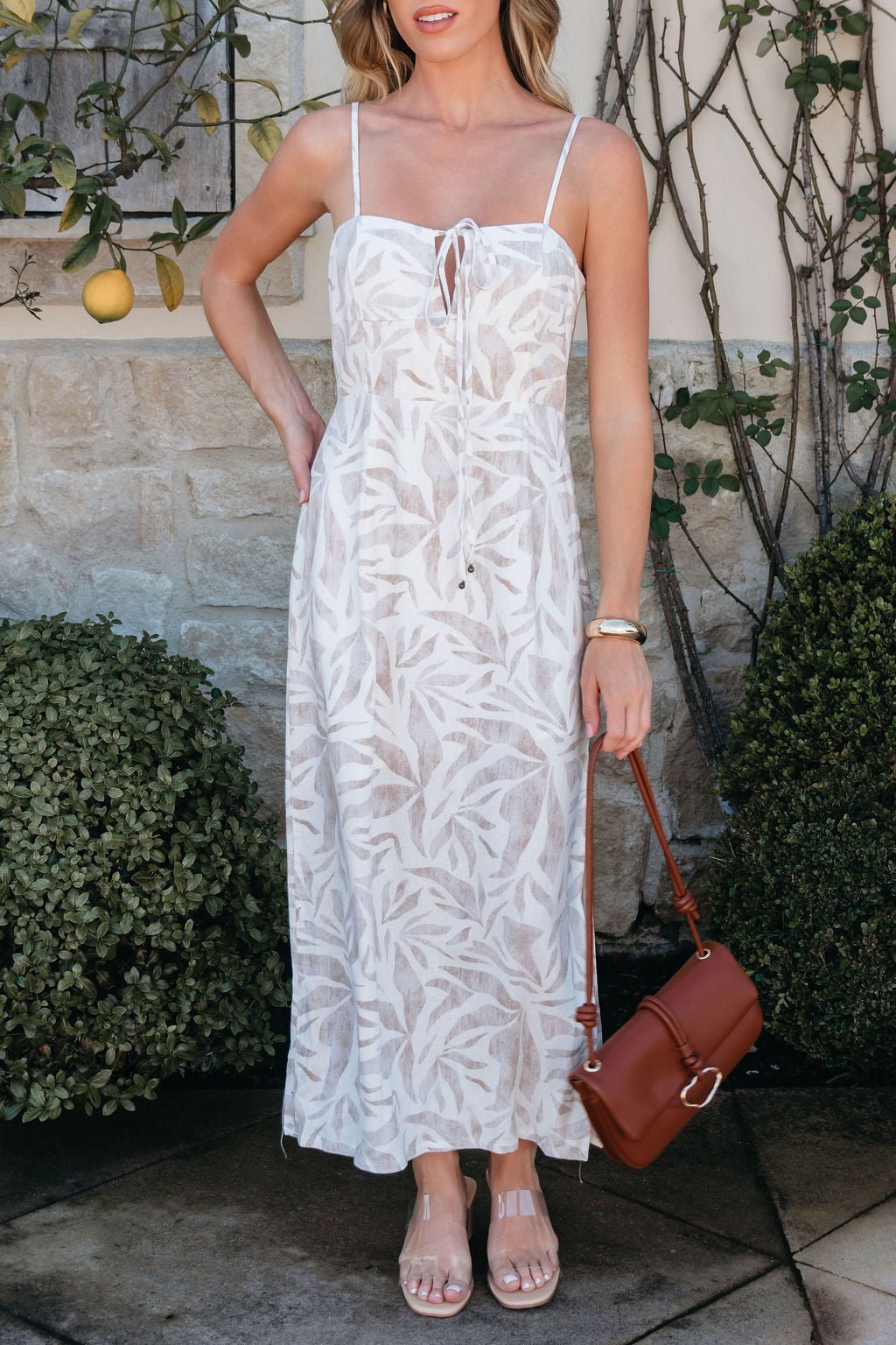 Woman in a Palm Vista White Maxi Dress stands by a stone wall and green bushes, holding a brown handbag.