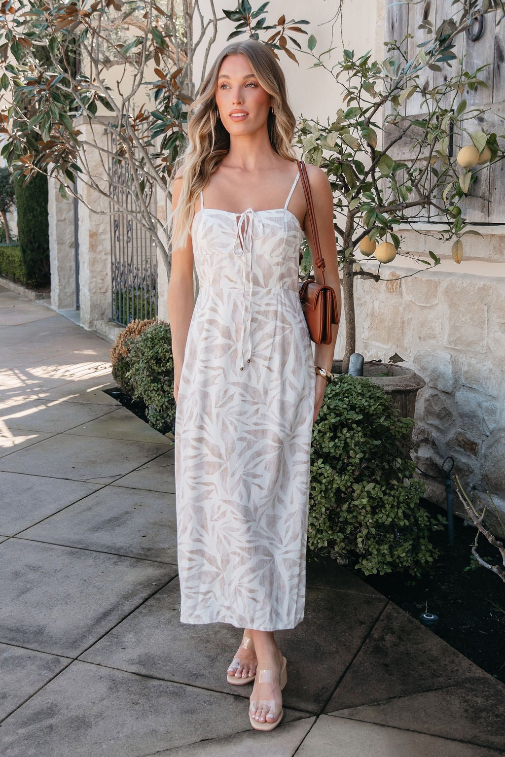 A woman in the Palm Vista White Maxi Dress and sandals stands by a stone wall, holding a brown shoulder bag.