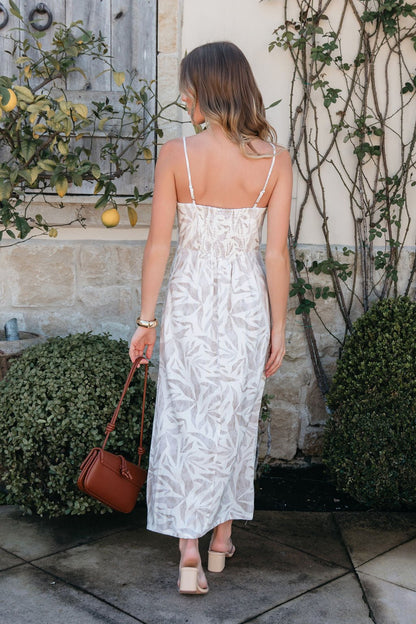 A woman in the Palm Vista White Maxi Dress with v-cut detail stands outside with a brown handbag amid greenery and stone walls.