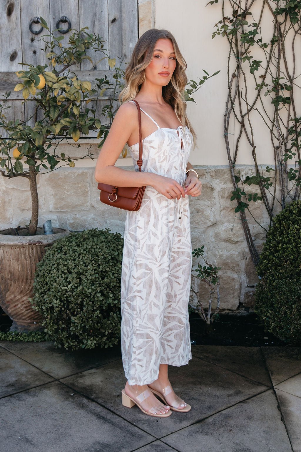 Woman in the Palm Vista White Maxi Dress with v-cut detail holds a brown shoulder bag by greenery and a stone wall.
