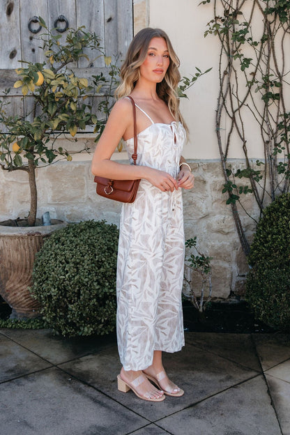 Woman in the Palm Vista White Maxi Dress with v-cut detail holds a brown shoulder bag by greenery and a stone wall.