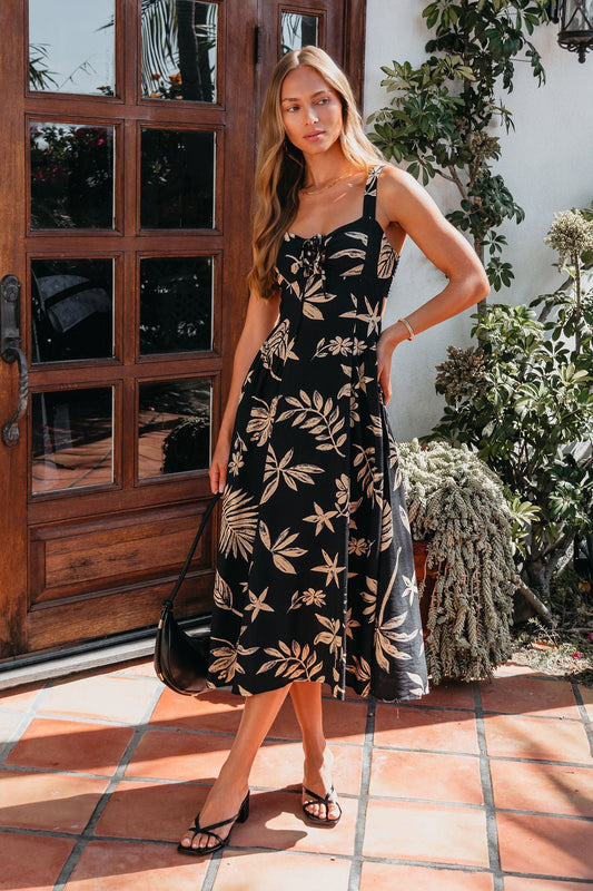 Woman in a Paloma Black Tropical Print Midi Dress stands by a wooden door, holding a black handbag.