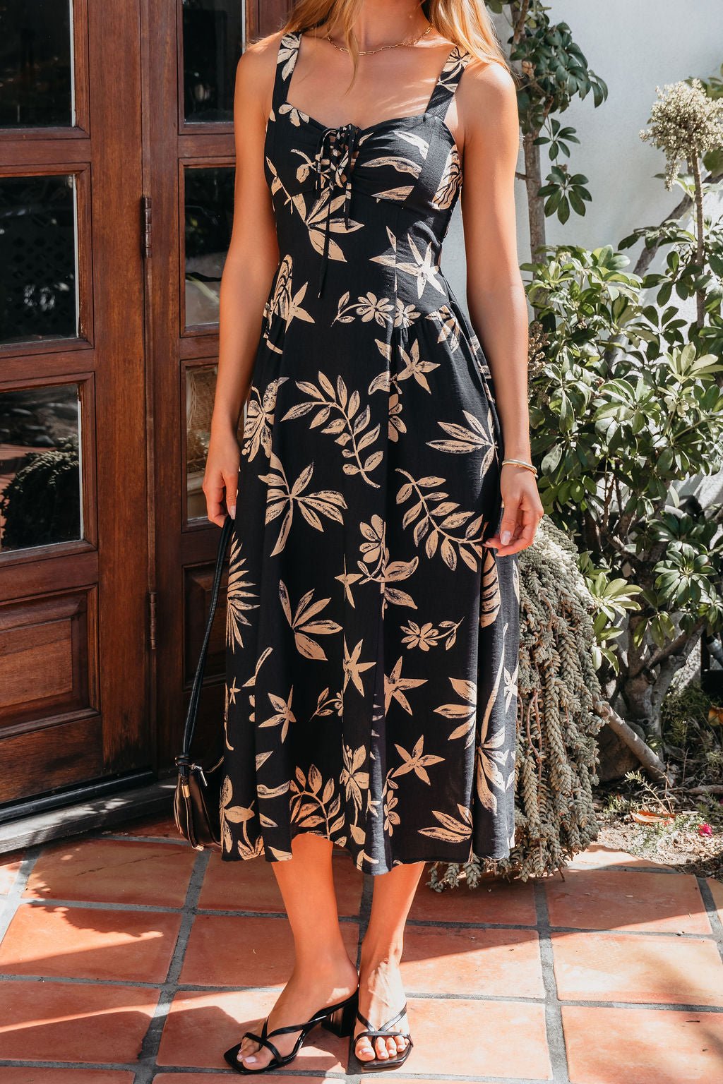 Woman in the Paloma Black Tropical Print Midi Dress stands on a tiled patio near wooden doors and greenery.