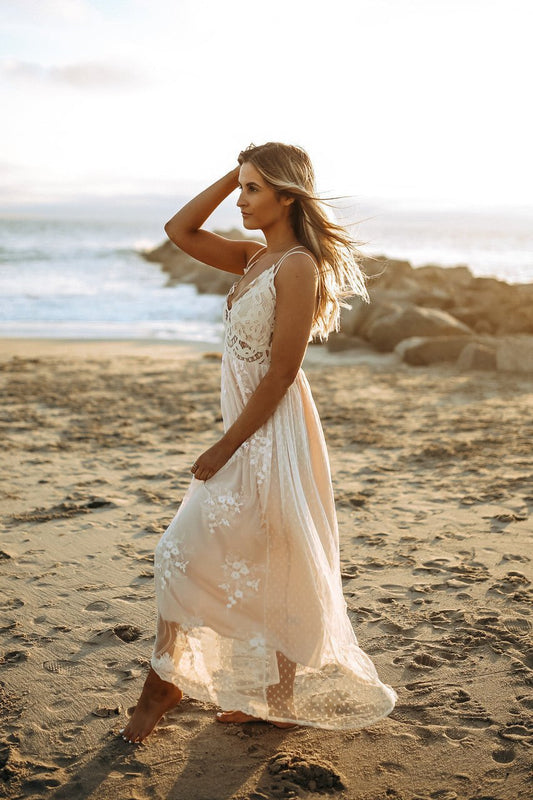 Woman in the Paradise Island Scalloped Vanilla Lace Maxi Dress walks barefoot on a sandy beach at sunset, wind in her hair.