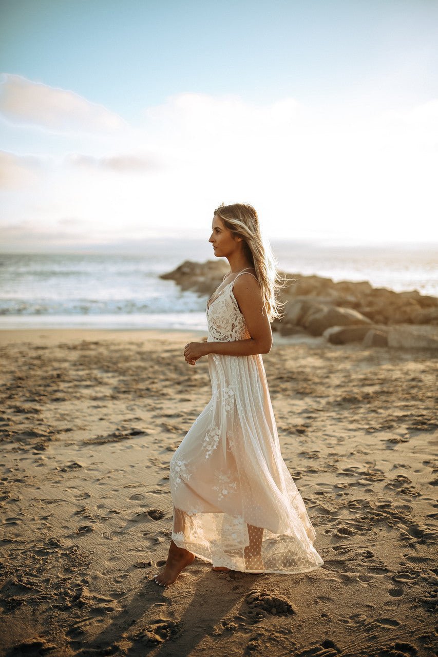 A woman in the Paradise Island Scalloped Vanilla Lace Maxi Dress walks barefoot on a sandy beach at sunset.