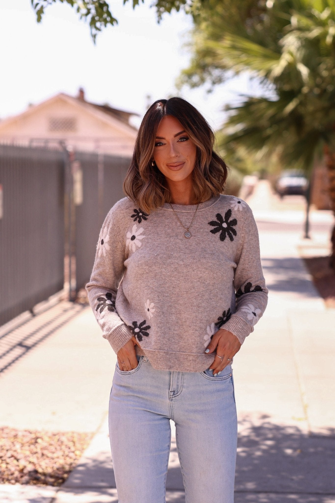 Woman stands on a sunny sidewalk, smiling in the Penelope Mocha Flower Detail Sweater, paired with light blue jeans.