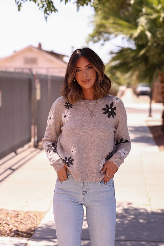 Woman stands on a sunny sidewalk, smiling in the Penelope Mocha Flower Detail Sweater, paired with light blue jeans.