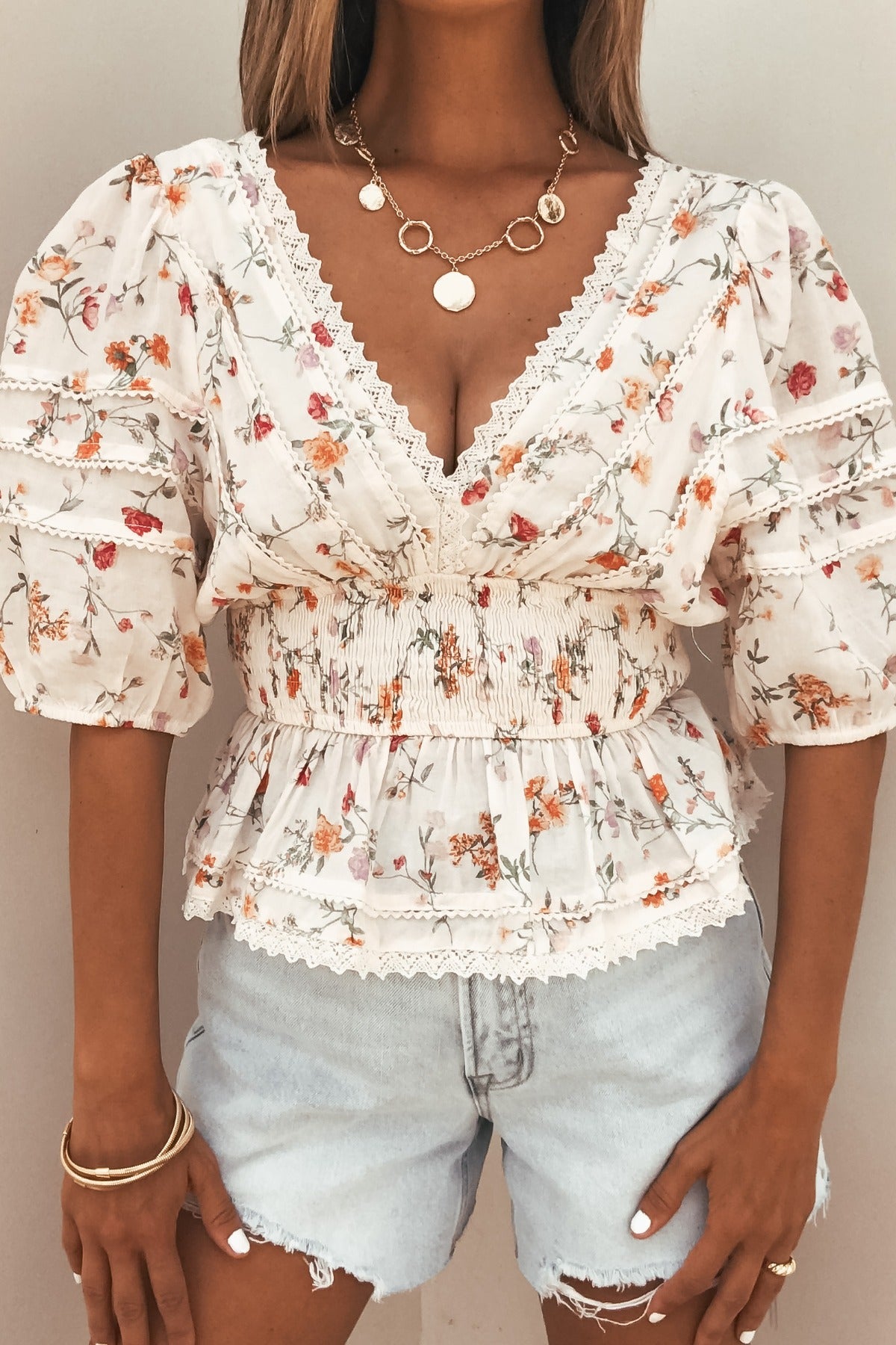 Woman wears the Petal Bloom Floral Smock Top - Cream with lace trim, puff sleeves, light denim shorts, and gold jewelry.