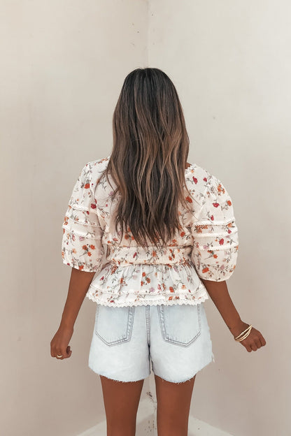 Woman with long wavy hair wears the Petal Bloom Floral Smock Top - Cream and light denim shorts, facing away from the camera.
