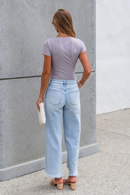 Woman with light brown hair in a lavender top and Petra High Rise Wide Leg Jeans - Light Wash stands facing a gray wall, holding a white clutch.