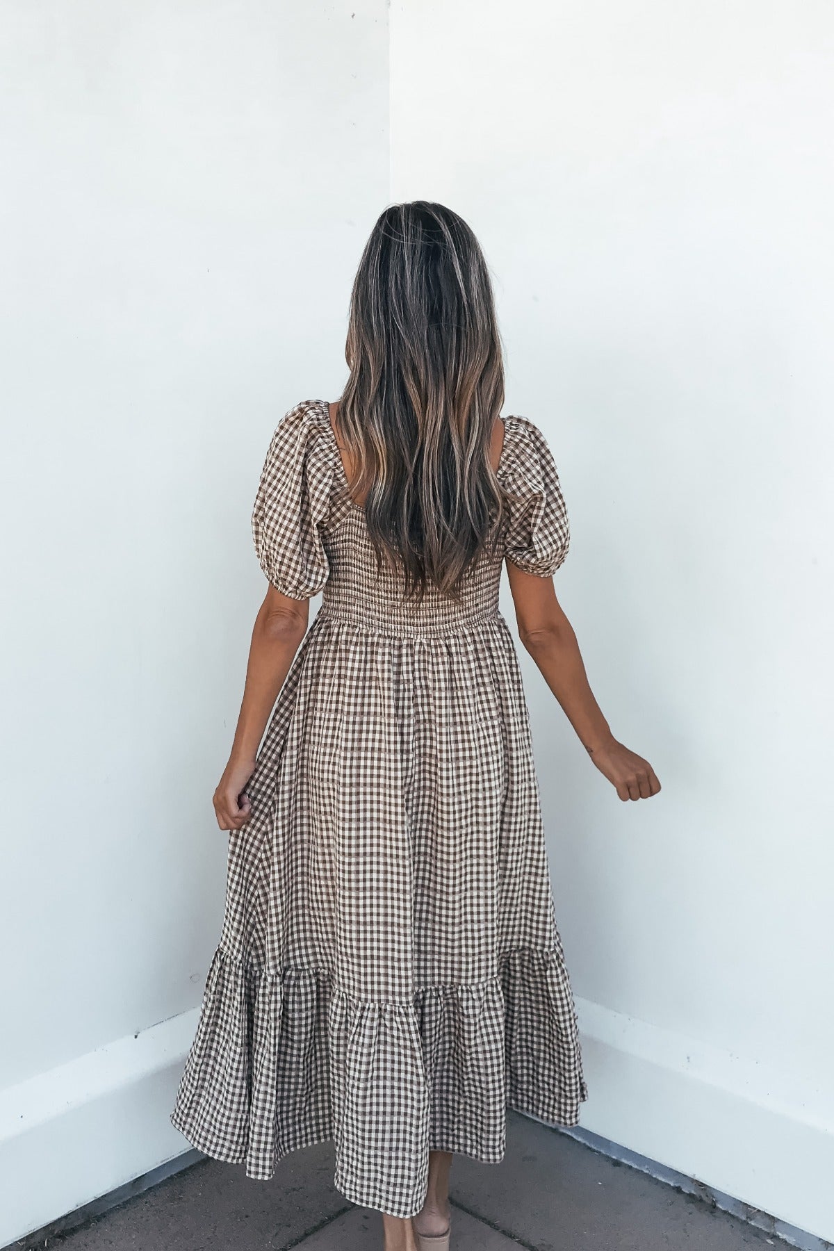 Woman in the Picnic Plaid Smock Midi Dress - Mocha faces a white corner, back to camera, on a tiled floor.