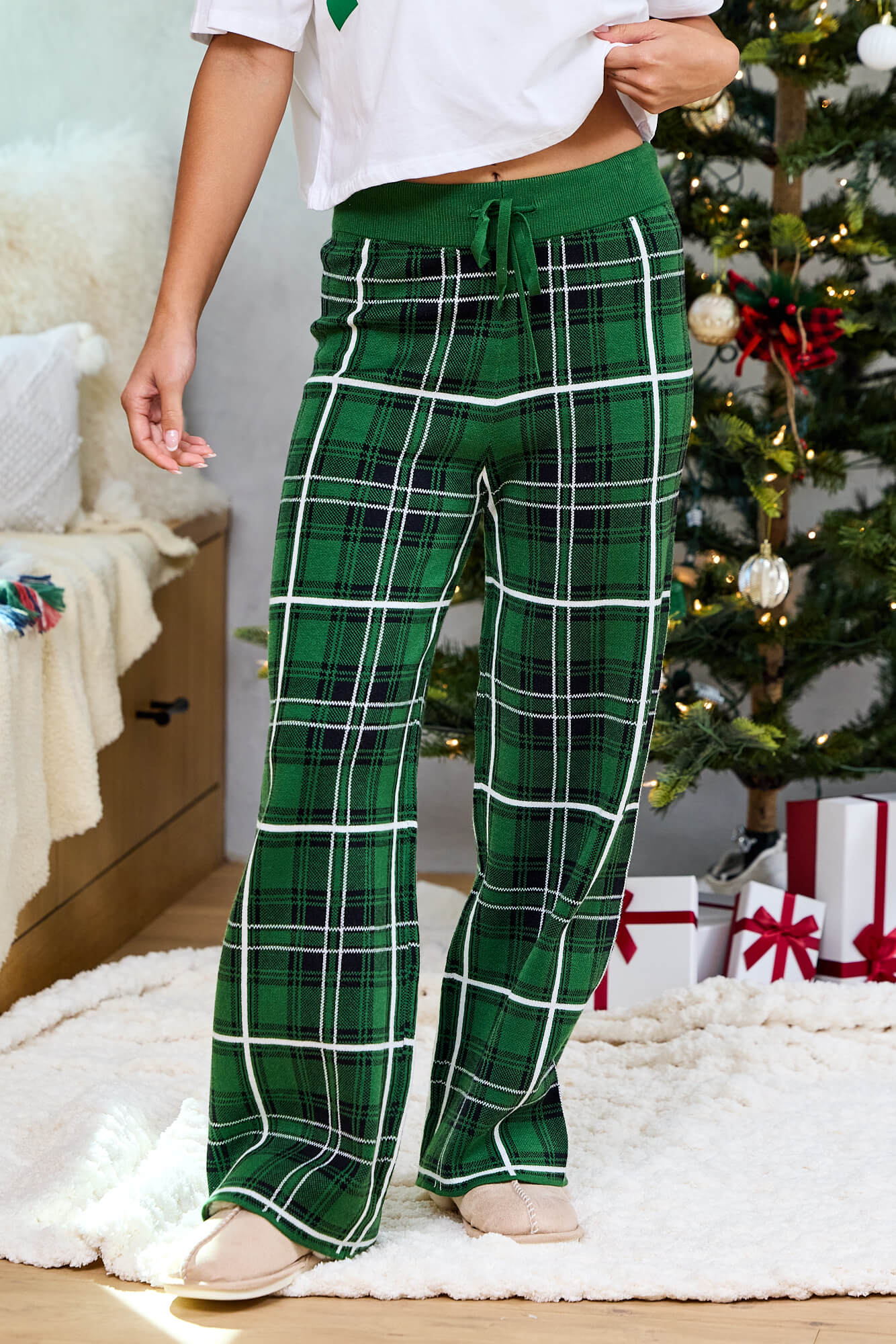Someone in Pine Mist Plaid Sweater Pajama Pants stands by a Christmas tree with presents and festive décor in the background.