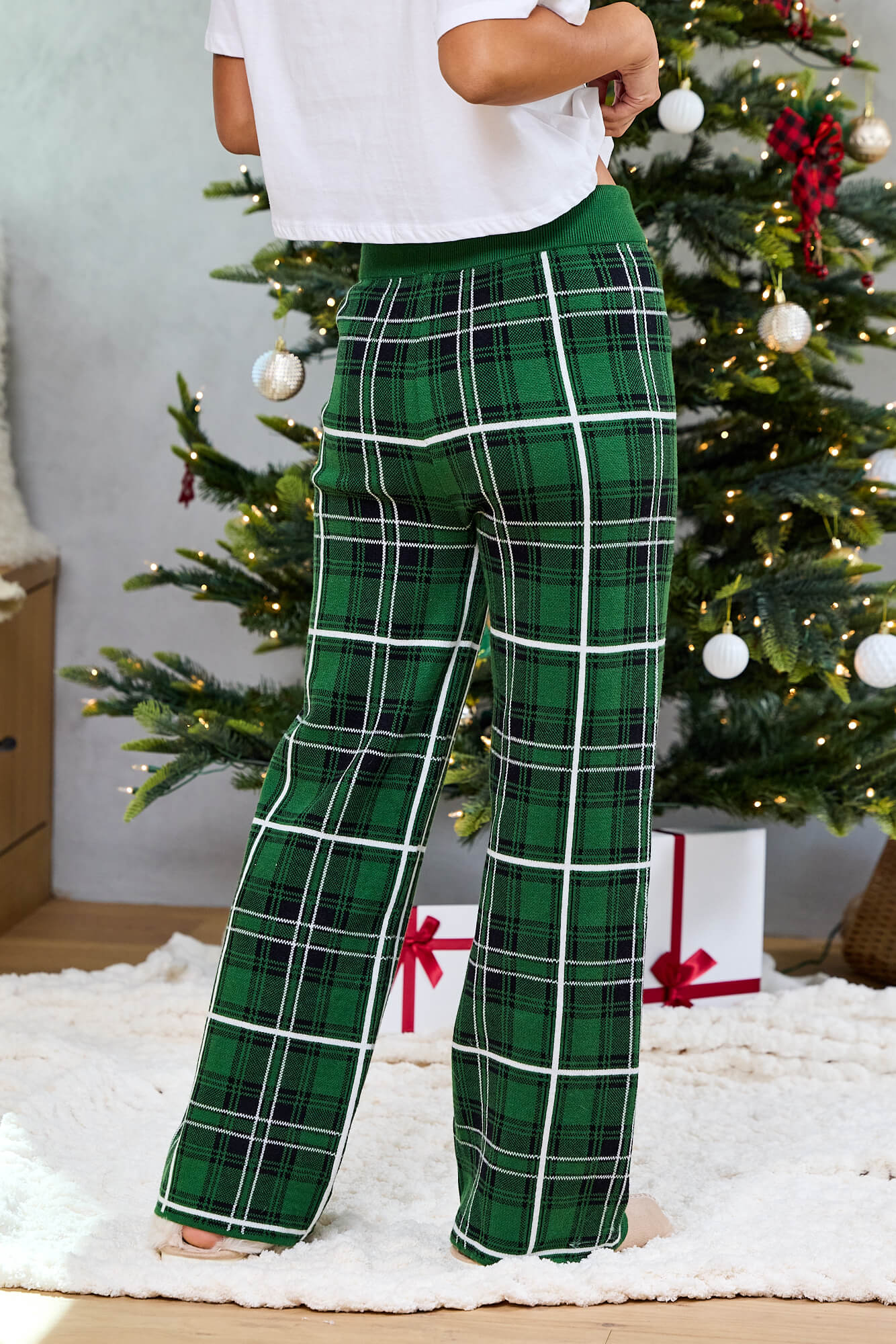 Person in Pine Mist Plaid Sweater Pajama Pants stands by a festive Christmas tree with wrapped gifts on a white rug.