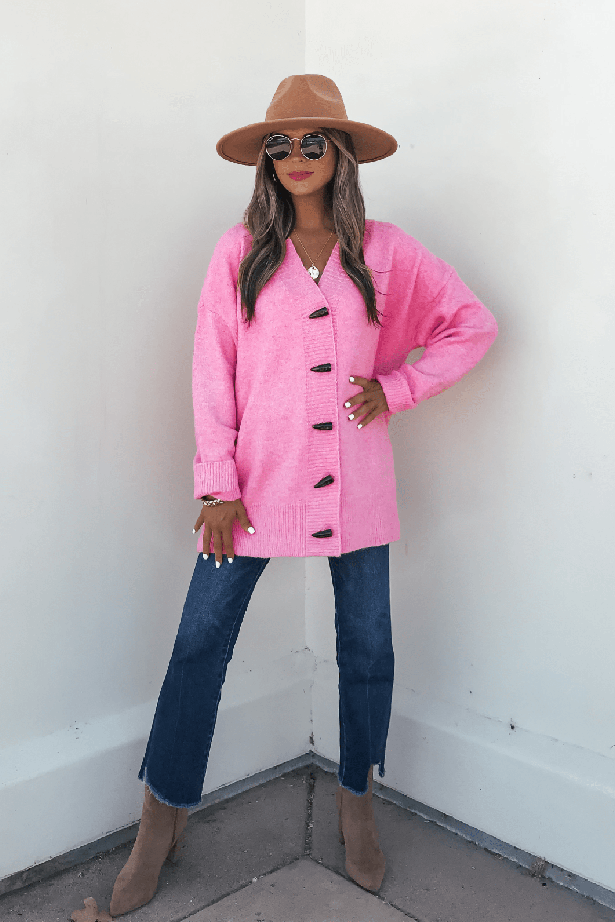 A woman styles the Pink Brushed Button Down Sweater Cardigan with blue jeans and boots against a white wall for a chic fall look.