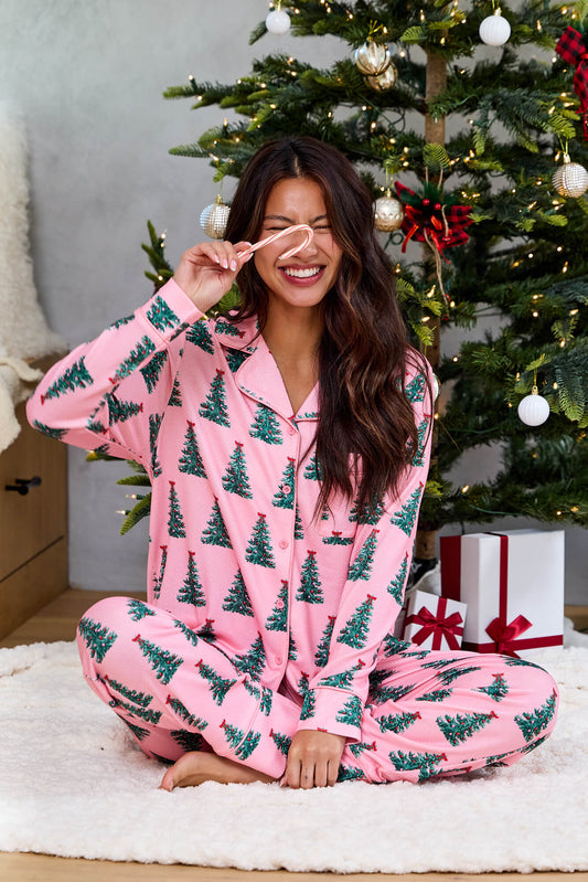 A smiling woman in a Pink Christmas Tree Print Pajama Set sits by a decorated tree, holding a candy cane over one eye.