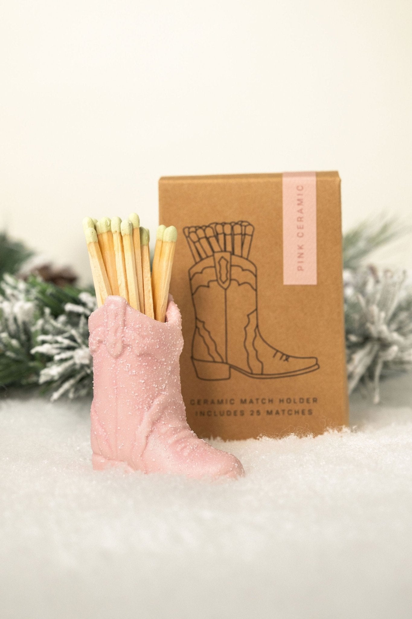 The Pink Cowboy Boot Match Holder sits in snow with a brown box and pine—fun western decor and a great gift idea.