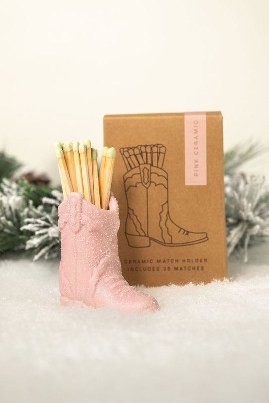 The Pink Cowboy Boot Match Holder sits in snow with a brown box and pine—fun western decor and a great gift idea.