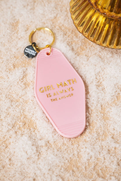The Pink Girl Math Motel Keychain features gold "Girl Math is always the answer" text and rests on sandy texture by a ribbed amber glass.