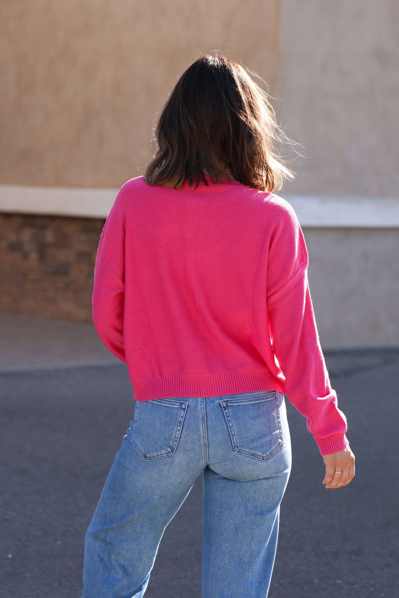 A person with shoulder-length brown hair wears a Pink Henley Button Up Sweater and blue jeans while walking outdoors, seen from behind.