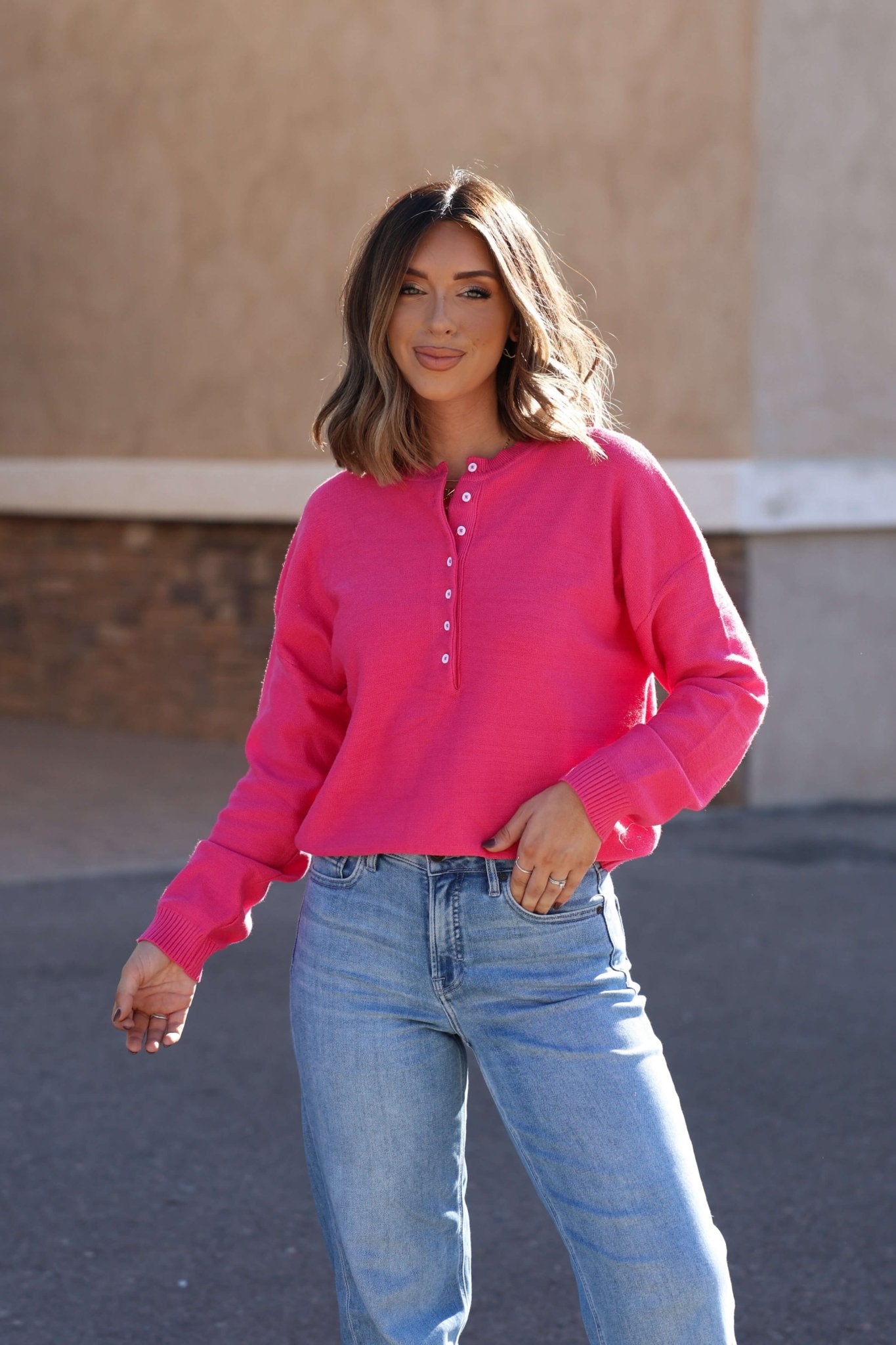 Smiling outdoors, she wears the Pink Henley Button Up Sweater with light blue jeans—your must-have fall essential.