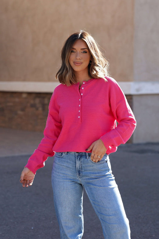 Smiling outdoors, she wears the Pink Henley Button Up Sweater with light blue jeans—your must-have fall essential.