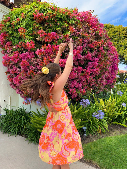 A woman in a Pink Paisley Floral Print Mini Dress dances joyfully before blooming pink bougainvillea in a sunlit garden.