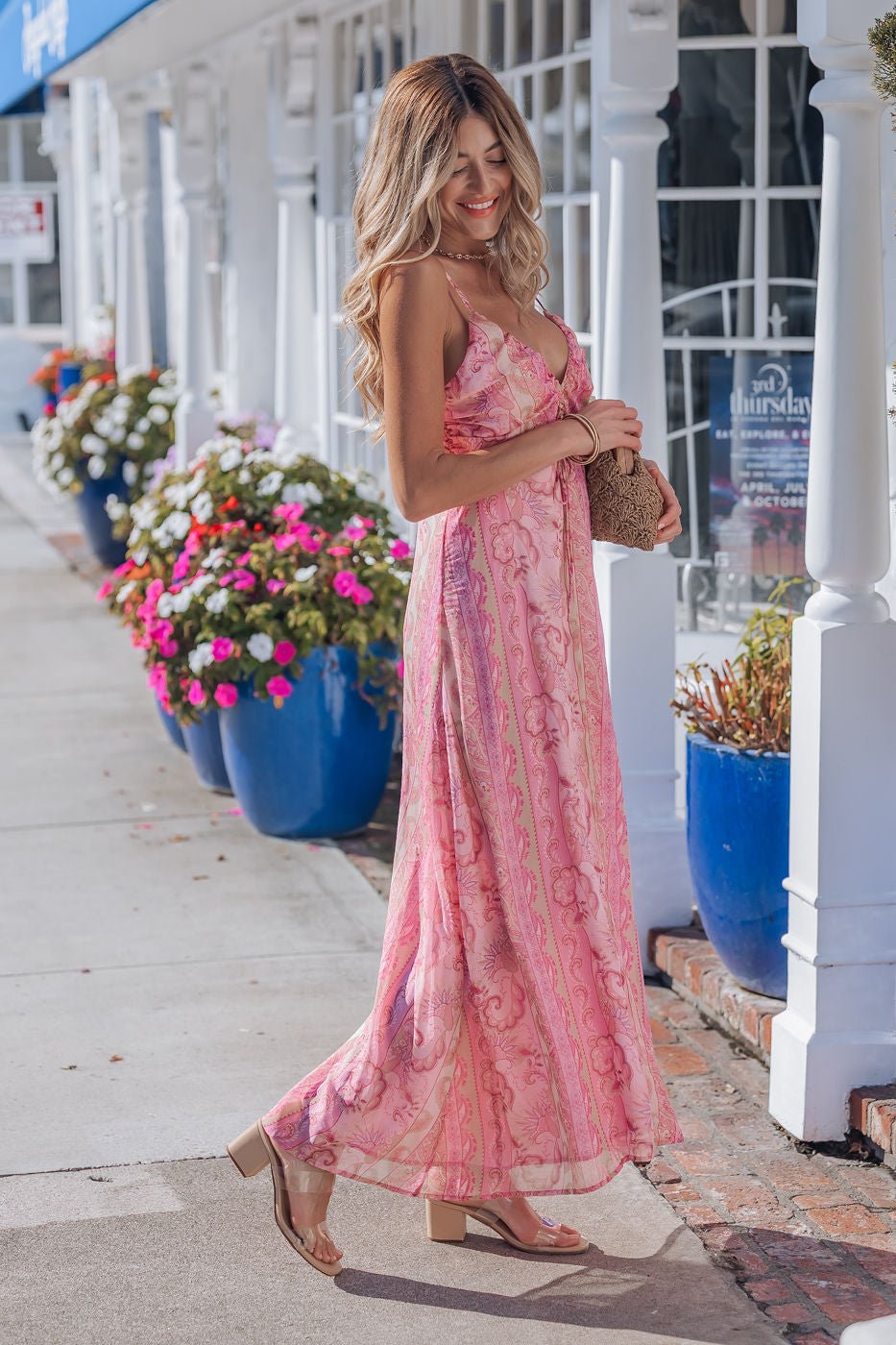 A woman in a Pink Paisley Print Maxi Dress and sandals smiles, strolling past blue flower pots with a small brown bag.