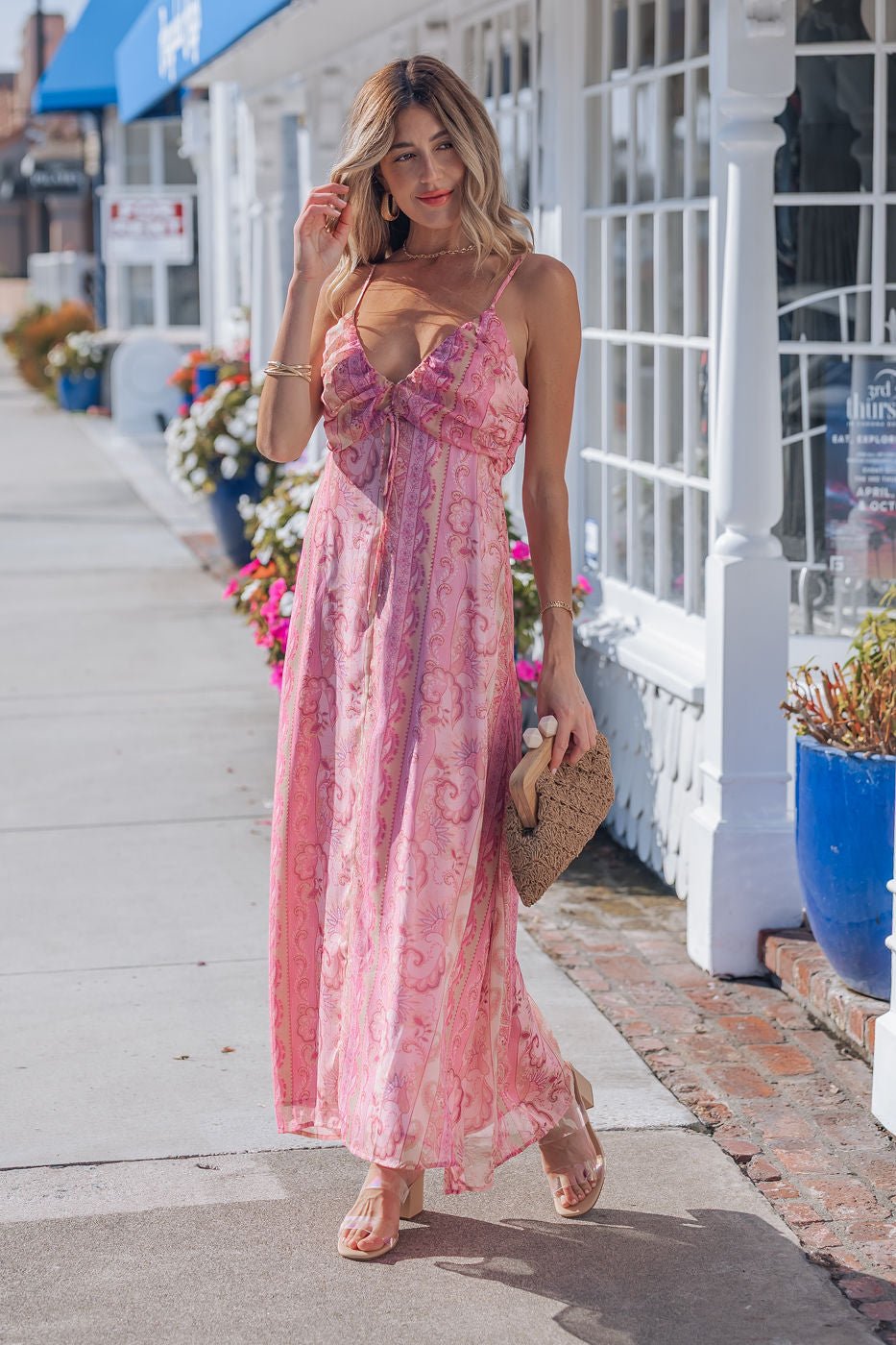 Woman wears a Pink Paisley Print Maxi Dress with adjustable straps, holding a woven clutch by a flower-filled white storefront.