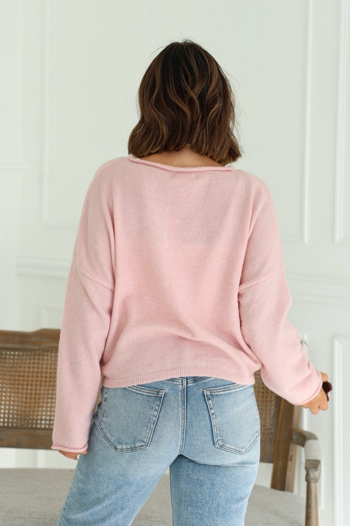A woman wears the Pink Rolled Boat Neck Sweater with blue jeans, facing away in a bright room, highlighting this chic layering piece.