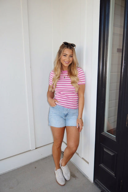 A woman in a Pink Short Sleeve Striped Sweater, denim shorts, and white sneakers smiles by a black door.