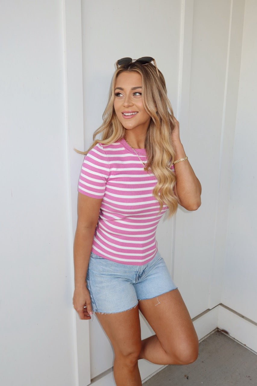 Woman with long wavy hair smiles by a white wall, wearing the Pink Short Sleeve Striped Sweater and denim shorts.