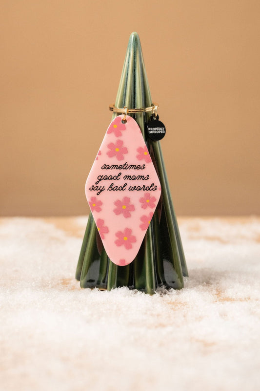 A fun gift: Pink Sometimes Good Moms Say Bad Words Motel Keychain, shown with flowers on a snowy ceramic tree.
