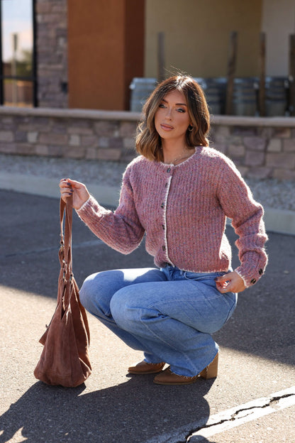 A woman wearing a Pink Speckled Button Down Cardigan and blue jeans squats outdoors, holding a brown bag and looking at the camera.